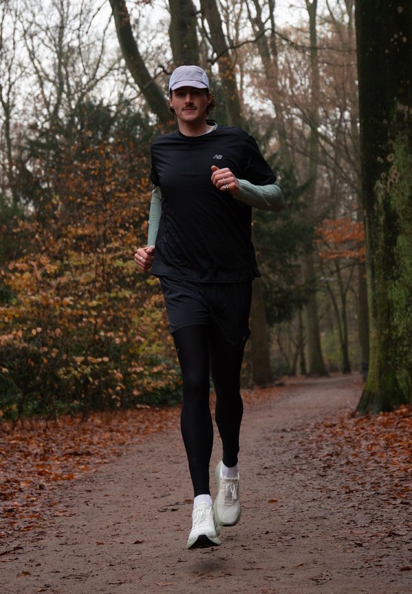 Mann läuft auf einem mit Blättern bedeckten Waldweg, trägt schwarze Sportbekleidung, weiße Kappe und weiße Laufschuhe im Herbst.