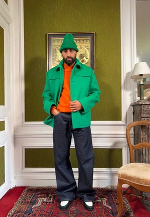 Hombre de pie en el interior, vistiendo un gorro de lana verde y chaqueta, suéter naranja, pantalones anchos oscuros, zapatos blancos y negros, en una habitación con paredes verdes y alfombra roja.