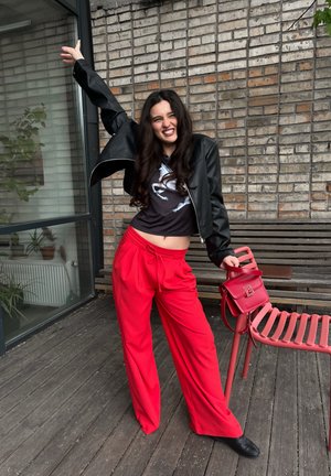 Chaqueta de cuero negra corta, camiseta gráfica negra, pantalones rojos holgados con cordón y un pequeño bolso rojo. Suelo de madera y pared de ladrillo de fondo.