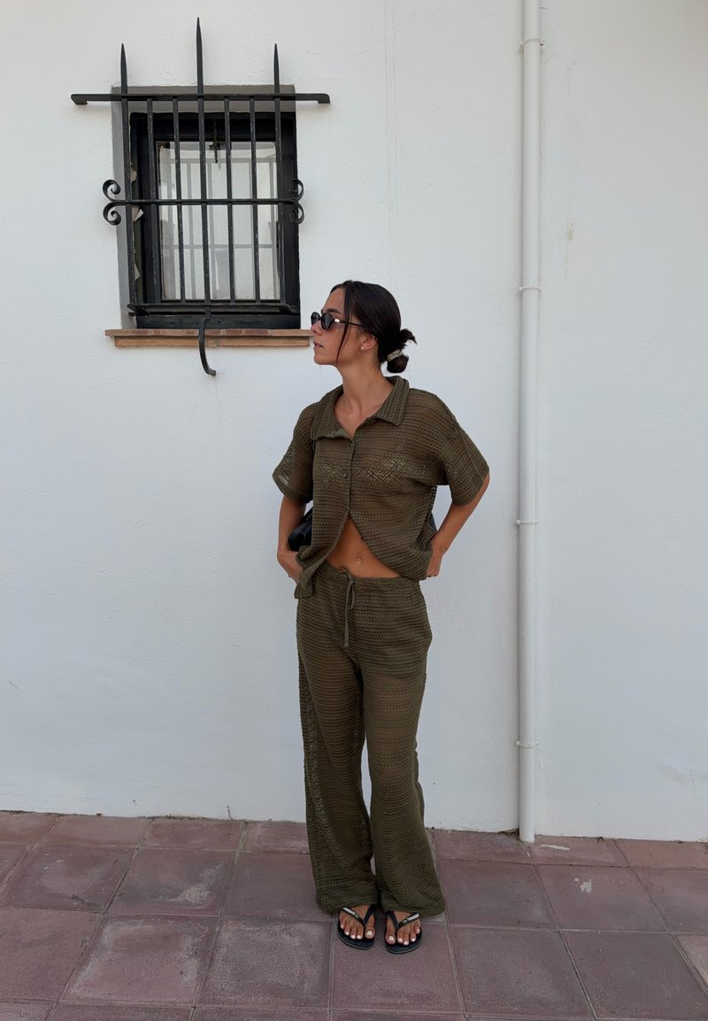 Γυναίκα με σκούρο πράσινο πλεκτό τοπ και παντελόνια, με γυαλιά ηλίου και σανδάλια, στέκεται κοντά σε ένα λευκό τοίχο με παράθυρο με κάγκελα.