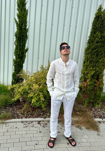 Camisa y pantalones de lino blancos, sandalias marrones, de pie cerca de plantas verdes, mirando hacia arriba, con una pared corrugada de color claro de fondo.