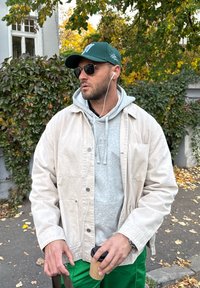 Béžová bunda přes šedou mikinu s kapucí, zelená baseballová čepice a sluneční brýle. Drží kelímek kávy, má zelené kalhoty a pozadí tvoří podzimní listí.