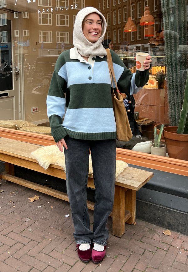 Chunky randig tröja i grön, ljusblå och mörkblå; grå jeans; vinröda tofflor; bär en krämfärgad stickad huva; håller i en kaffekopp.