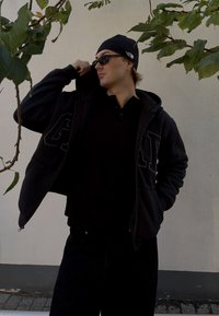 Čierna mikina na zips s logom, podšitá interiérom, spárovaná s čiernym polo tričkom a tmavými nohavicami. Doplnky zahŕňajú čierne slnečné okuliare a čiapku.