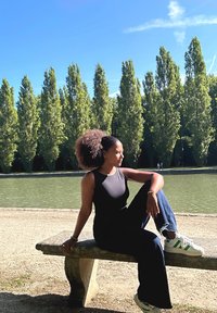 Schwarzes ärmelloses Oberteil, hoch taillierte dunkle Jeans, weiße Sneakers mit grünen Akzenten. Sitzt auf einer Steinbank am Wasser mit Bäumen im Hintergrund.