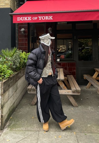 Chaqueta acolchada negra, top corto de estampado de leopardo, pantalones negros holgados con rayas blancas, botas color marrón y gorra con estampado de leopardo. Entorno al aire libre.