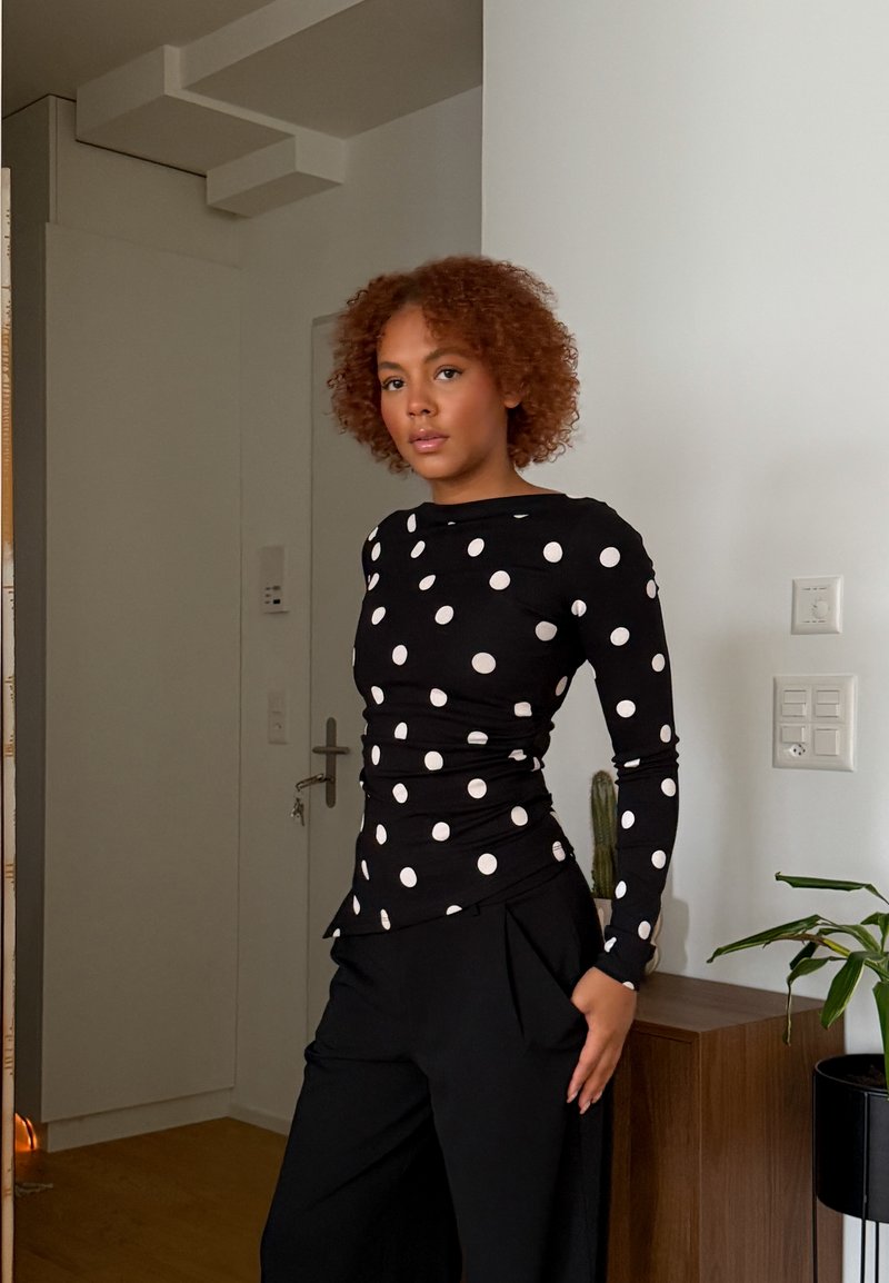 Joven con el cabello rizado, vestida con una blusa negra de manga larga con lunares blancos y pantalones negros, de pie en el interior cerca de una planta y una puerta.