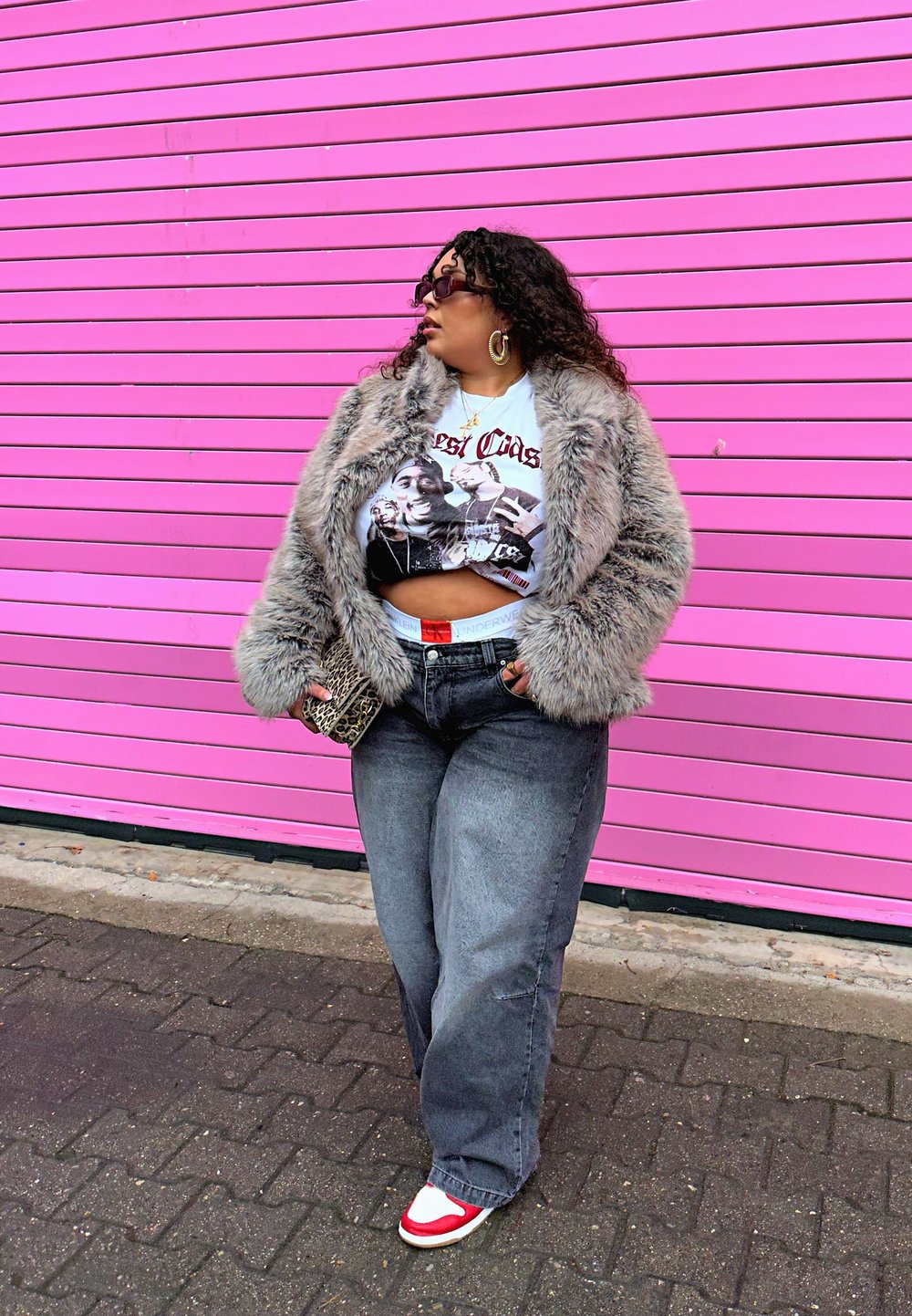 Cappotto grigio in pelliccia sintetica, t-shirt crop bianca con grafica, jeans scuri a vita alta, sneakers rosse e bianche, occhiali da sole ovali, pochette con stampa animale, sfondo rosa.