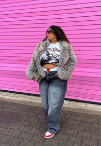 Cappotto grigio in pelliccia sintetica, t-shirt crop bianca con grafica, jeans scuri a vita alta, sneakers rosse e bianche, occhiali da sole ovali, pochette con stampa animale, sfondo rosa.