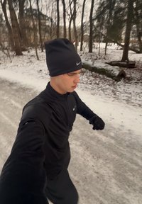 Jeune homme vêtu de noir courant sur un sentier enneigé dans une forêt d'hiver aux arbres dénudés.