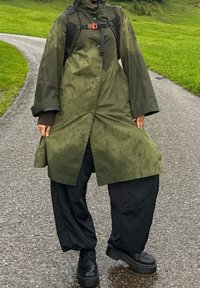 Grønn vanntett regnjakke med løs passform, lange ermer og høy krage, kombinert med svarte oversized bukser og kraftige svarte vanntette støvler.