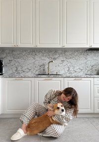 Femme en pyjama à carreaux assise sur le sol de la cuisine, embrassant un chien corgi marron et blanc près des armoires blanches et du dosseret en marbre.