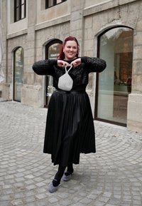 Μαύρη πλισέ φούστα, λαμπερό μαύρο κοντό πουλόβερ, γκρι τσάντα με υφή, παπούτσια με σχέδιο μαύρο και άσπρο, γυναίκα που κρατά την τσάντα μπροστά της.