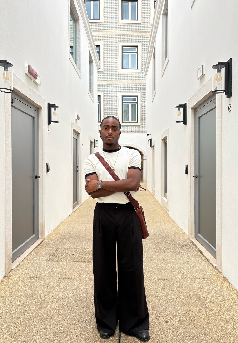 Refleksiv sølvklokke, beige kortermet skjorte med svarte detaljer, svarte vide bukser, og brun skinnveske mot en lys korridor.