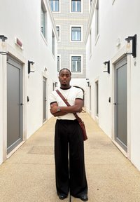 Refleksiv sølvklokke, beige kortermet skjorte med svarte detaljer, svarte vide bukser, og brun skinnveske mot en lys korridor.