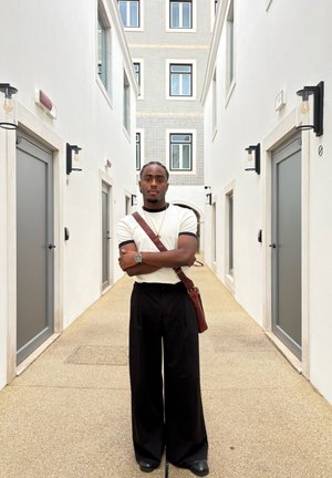 Reloj plateado reflectante, camisa de manga corta beige con acentos negros, pantalones de pierna ancha negros y bolsa de cuero marrón en un pasillo claro.