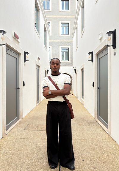 Reloj plateado reflectante, camisa de manga corta beige con acentos negros, pantalones de pierna ancha negros y bolsa de cuero marrón en un pasillo claro.