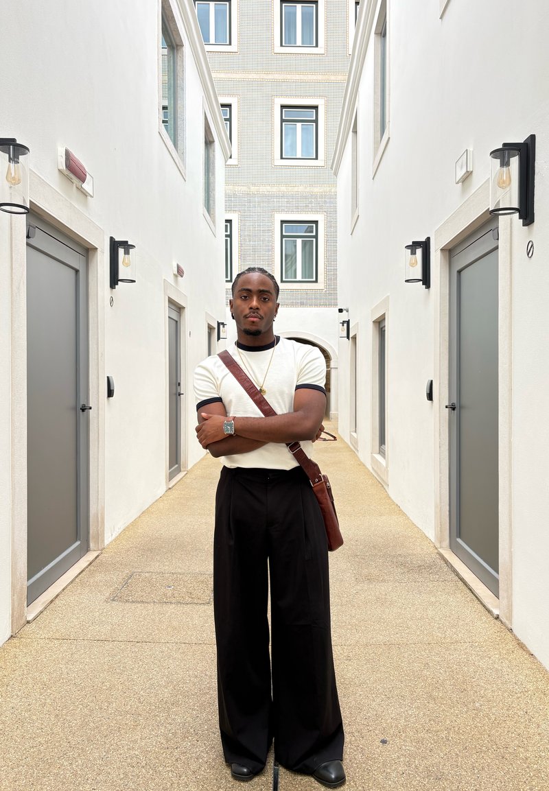 Peegeldav hõbedane kell, beež lühikeste varrukatega särk mustade detailidega, mustad laiad püksid ja pruun nahkrahakott heleda koridori taustal.