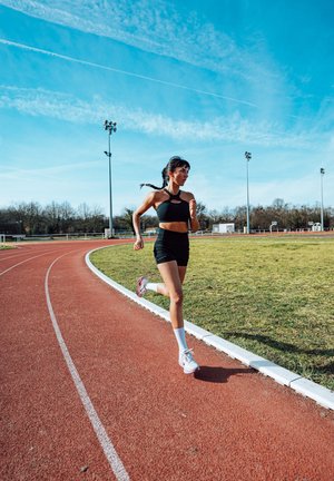 Mujer con ropa deportiva negra corriendo en una pista exterior roja con campo de césped bajo un cielo azul despejado durante el día.