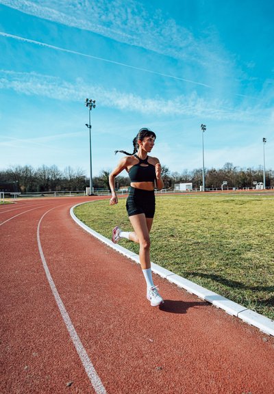 Mujer con ropa deportiva negra corriendo en una pista exterior roja con campo de césped bajo un cielo azul despejado durante el día.