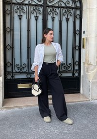 Witte linnen shirt, groene tanktop, wijde zwarte broek, grijze schoenen, houdt een zwart en crèmekleurige handtas vast, tegen een sierlijke zwarte deur.