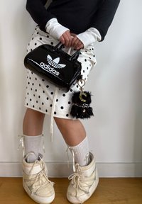 Čierna lesklá kabelka Adidas s bielym logom, biela sukňa s bodkami, béžové čižmy a nadýchaný čierny prívesok na kľúče so zameraním na písmeno.