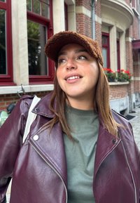 Maroon læder bikerjakke med sølv lynlåsdetaljer, olivengrøn t-shirt og brun struktureret kasket, sat mod røde vinduer og murstensvæg.