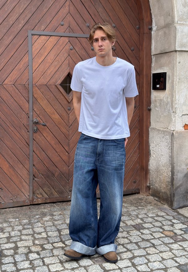 Jeune homme aux cheveux clairs se tient dans une rue pavée, portant un t-shirt blanc, un jean large et ample, et des chaussures marron devant une grande porte en bois.
