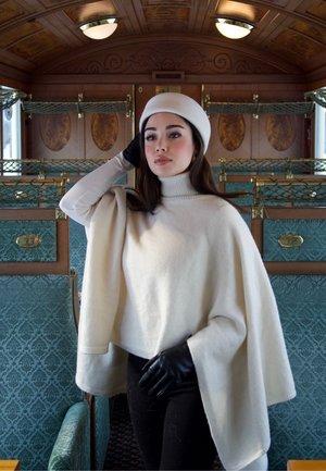 Joven mujer con boina blanca, capa color crema y guantes negros, de pie dentro de un vagón de tren vintage con asientos estampados en color verde azulado.