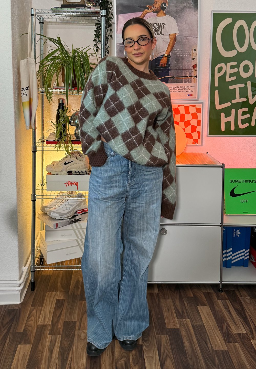 Übergroßer brauner und türkisfarbener Argyle-Pullover, kombiniert mit hellblauen weiten Jeans. Schwarze Schuhe runden das Outfit ab. Zeitgenössisches Setting mit Regalen.