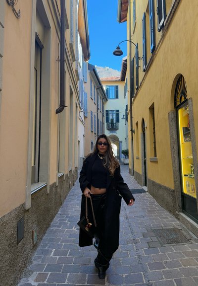Abrigo largo negro sobre una camiseta corta negra, combinado con pantalones negros y botas de tobillo. Callejón urbano con edificios amarillos y beige, cielo soleado.