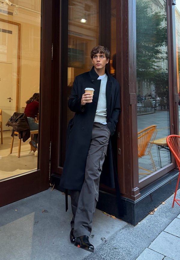 Black overcoat, grey jumper, and loose grey trousers. Holding a coffee cup; shiny black shoes. Wooden and glass café backdrop with orange chairs.