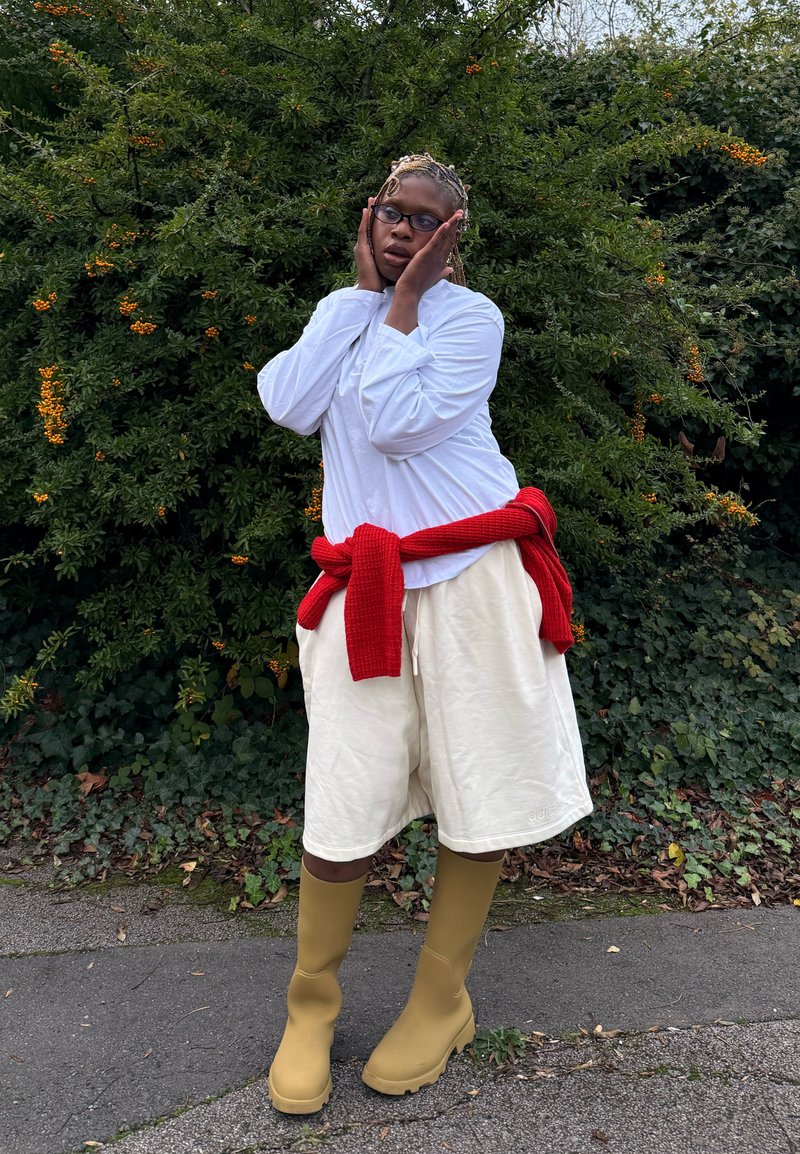 Camicia bianca a maniche lunghe, pantaloncini color crema, maglione rosso legato in vita e stivali di gomma gialli fino al ginocchio contro un folto fogliame verde.