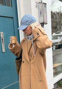 Femme en manteau camel et casquette bleue tenant une tasse de café, debout près d'une porte bleue portant le numéro 31 dans un cadre urbain.