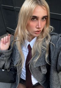 Blazer oversize in pied de poule grigio, camicia bianca e cravatta color bordeaux. Capelli biondi e lunghi, tiene in mano una piccola borsa nera, su uno sfondo scuro.