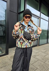 Pullover de polar multicolor com padrões geométricos, gola azul clara, combinado com calças pretas largas e uma mala de cintura preta.