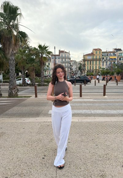 Joven mujer con pantalones blancos y top marrón usando teléfono con auriculares, caminando por la acera de una ciudad cerca de palmeras y edificios.