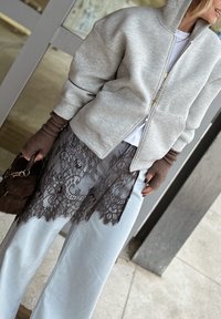 Femme portant une veste grise zippée, un tablier en dentelle marron par-dessus un jean bleu clair, des gants sans doigts marron, et tenant un sac à main marron foncé.