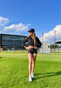 Crna kratka jakna, crne šortse s uzorkom i bijele tenisice s ljubičastim detaljima. U pozadini travnata površina i sportski objekt.