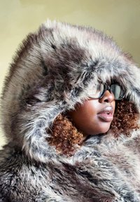 Kabát s kapucňou z umelých kožúškov v odtieňoch šedej a hnedej, s objemnou kapucňou a príjemnou textúrou. Číre okuliare nosené cez kučeravé vlasy.