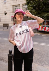 Šviesiai rožinė dryžuota per didelė marškinė su "NIKE" logotipu, derinama su juodomis kelnėmis ir rožine kepure, papildyta petine krepše ir kaklo papuošalu.