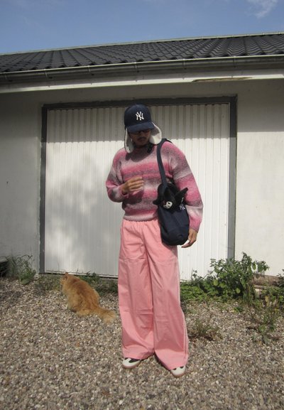 Pantalones de pierna ancha rosa, un suéter a rayas rosa y gris, y un sombrero azul marino. Un bolso negro con un animal de peluche. Un gato está en el fondo.