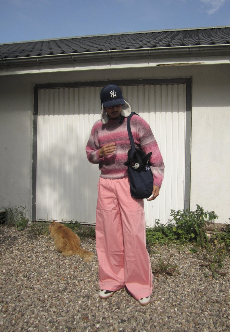 Pantalon large rose, un pull rayé rose et gris, et un chapeau bleu marine. Un sac noir avec un animal en peluche. Un chat est en arrière-plan.