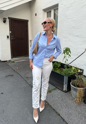 Camisa de botones a rayas azul claro, jeans blancos bordados con recortes florales y tacones de punta crema. Bolso marrón claro sobre el hombro.