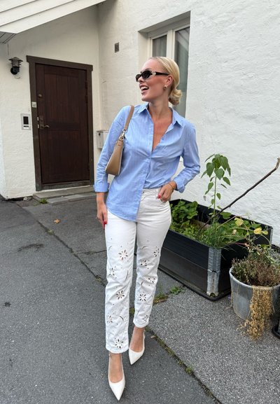 Camisa de botones a rayas azul claro, jeans blancos bordados con recortes florales y tacones de punta crema. Bolso marrón claro sobre el hombro.