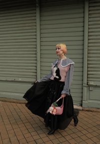 Strikkegenser med rosa base og svart kattemønster, rysjekrage; flytende svart skjørt; rosa håndveske med leketøycharmer; mønstret bakgrunn.