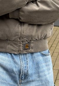 Γκρι bomber jacket με ribbed μανσέτες και τελείωμα, που διαθέτει μια καφέ λογότυπο τσεπάκι. Συνδυασμένο με ανοιχτόχρωμο μπλε τζιν παντελόνι, σε χαλαρή στάση.