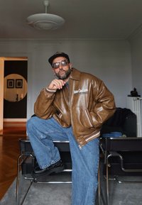 Mann som har på seg solbriller, svart caps, brun skinnjakke og blå jeans, sitter med ett ben på en svart stol i stuen.