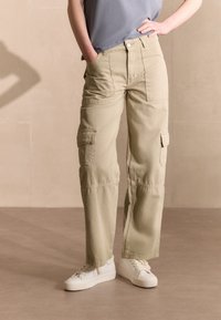 Pantaloni cargo beige chiaro con più tasche, gambe dritte e tessuto di cotone, abbinati a sneakers bianche e una maglietta blu-grigia.