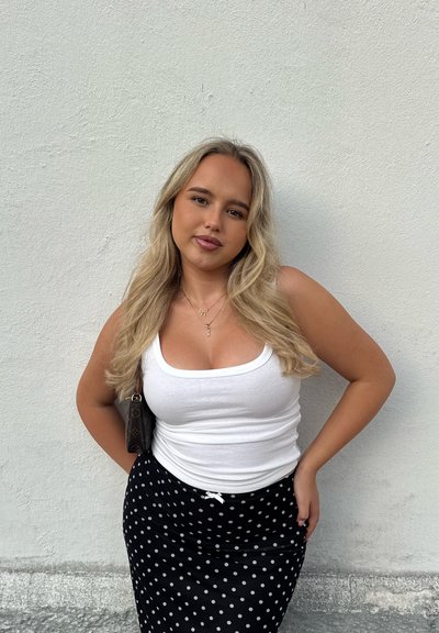 Top de tirantes blanco con textura acanalada combinado con una falda negra con lunares. Luce cabello rubio ondulado y lleva un bolso negro. Fondo de pared blanca lisa.